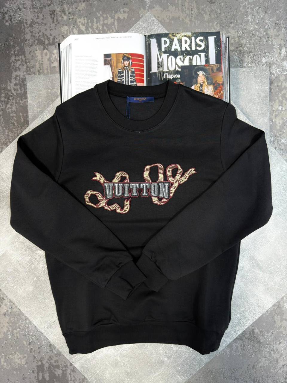 A black crewneck sweatshirt featuring a large Louis Vuitton monogram graphic across the chest in a snakeskin pattern with blue and silver lettering. The sweatshirt is displayed in a flat-lay arrangement on a gray textured surface, styled with an open magazine in the background.