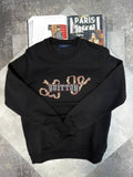 A black crewneck sweatshirt featuring a large Louis Vuitton monogram graphic across the chest in a snakeskin pattern with blue and silver lettering. The sweatshirt is displayed in a flat-lay arrangement on a gray textured surface, styled with an open magazine in the background.