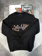 A black crewneck sweatshirt featuring a large Louis Vuitton monogram graphic across the chest in a snakeskin pattern with blue and silver lettering. The sweatshirt is displayed in a flat-lay arrangement on a gray textured surface, styled with an open magazine in the background.