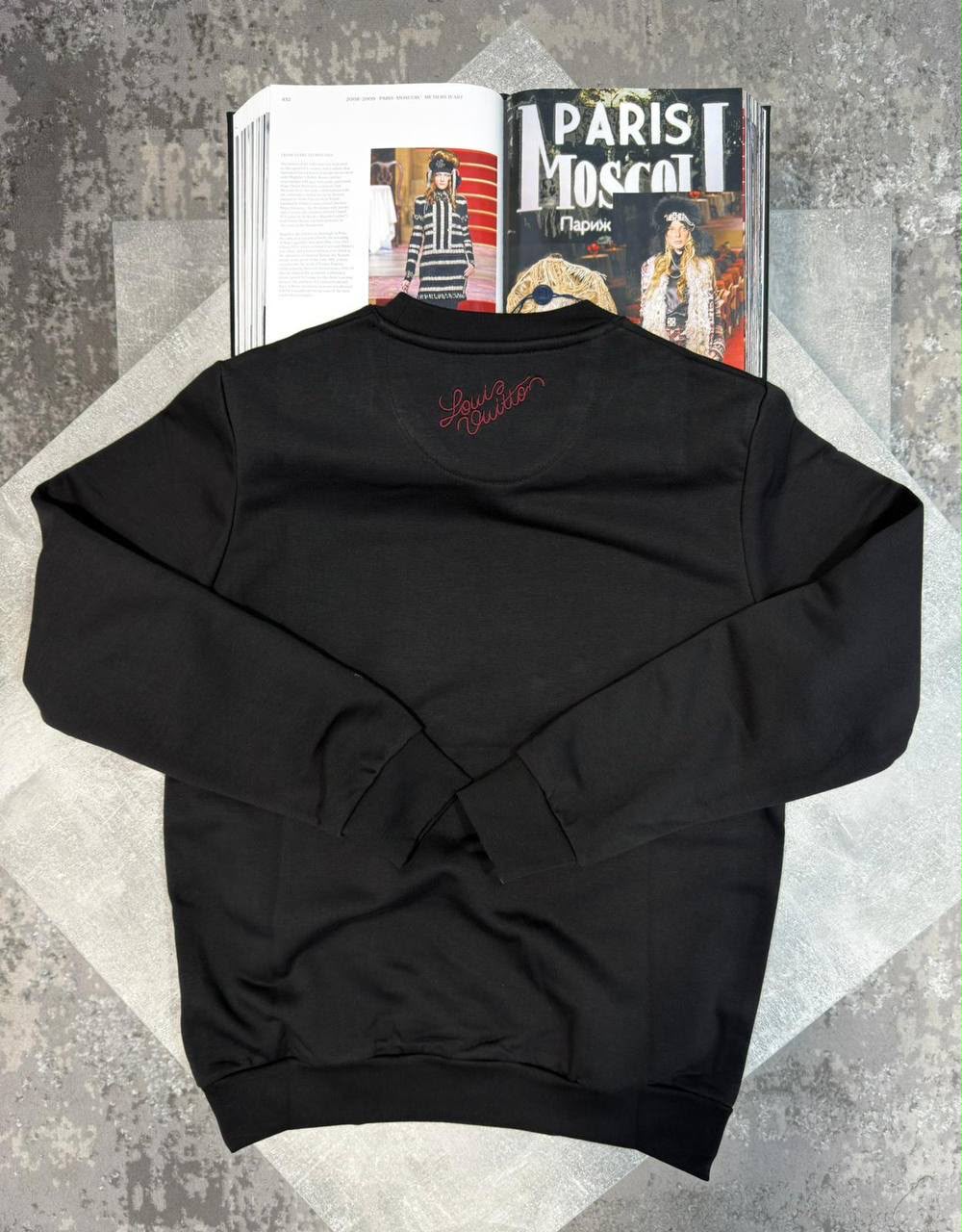 A black crewneck sweatshirt with a red embroidered script logo on the chest, laid flat on a gray patterned surface with open magazines behind it.