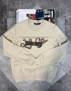 A cream-colored Louis Vuitton crewneck sweatshirt with a graphic print of a snake coiling around the brand's name, displayed on a textured gray surface with magazines in the background.