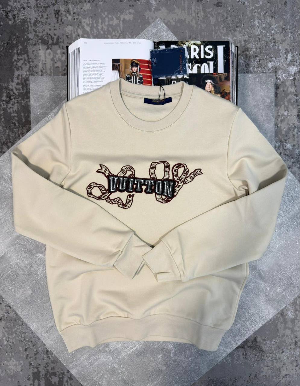 A cream-colored Louis Vuitton crewneck sweatshirt with a graphic print of a snake coiling around the brand's name, displayed on a textured gray surface with magazines in the background.