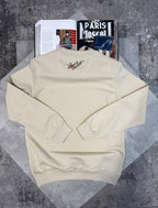 A cream-colored crewneck sweatshirt with a small embroidered logo on the back, laid flat on a textured gray surface with an open magazine underneath.