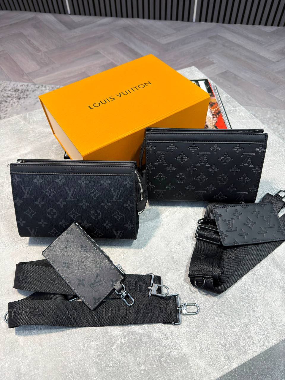 Louis Vuitton black monogram canvas wallet set with matching black monogram shoulder strap and a bright orange Louis Vuitton box on a light gray floor.