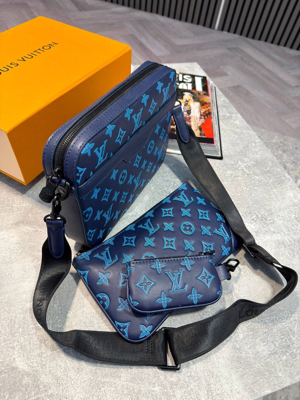 Louis Vuitton monogram shoulder bag in navy blue with a bright blue monogram pattern, shown with its matching pouch and orange Louis Vuitton box on a gray surface.