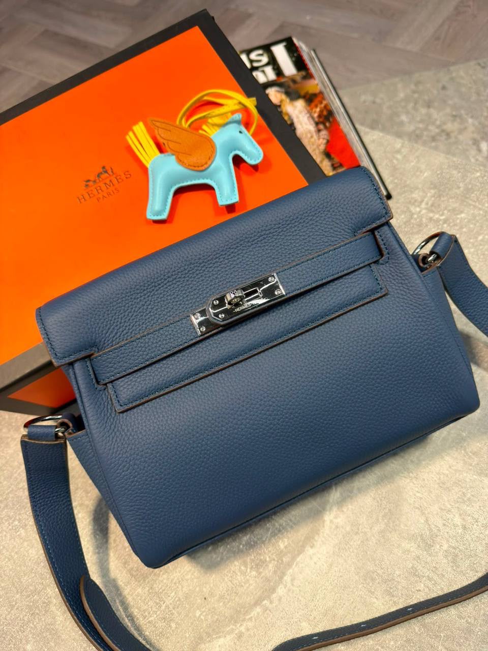 A blue pebbled leather handbag with silver hardware, displayed on a light gray stone surface next to an orange Hermès box and a small horse-shaped accessory.