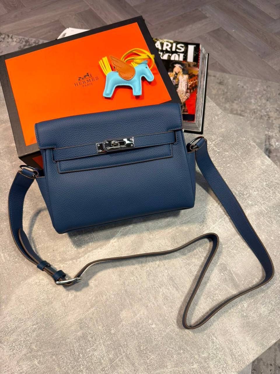 A blue leather crossbody bag with silver hardware, displayed on a gray floor next to its orange and black Hermès box and a magazine.