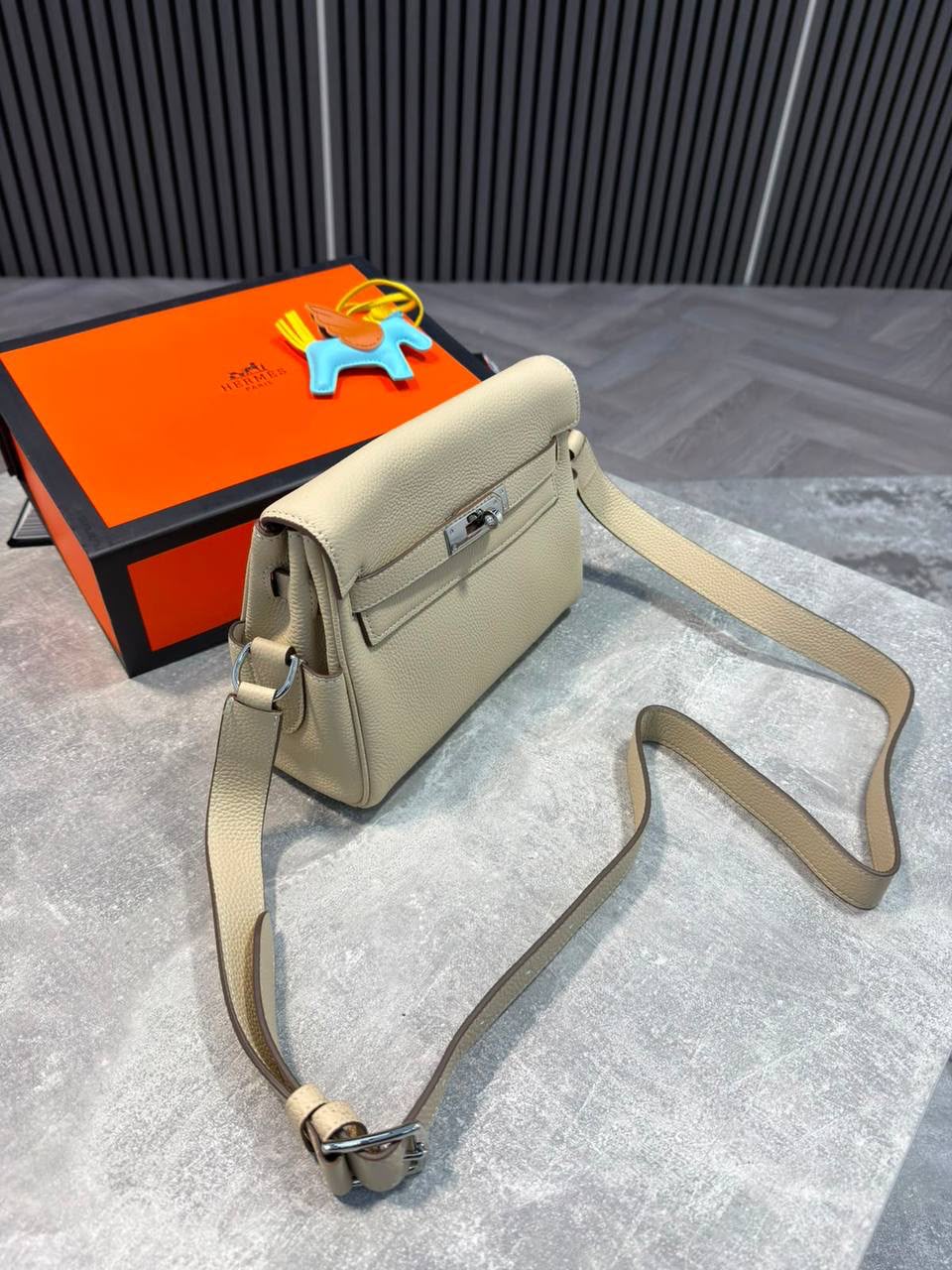 A beige leather shoulder bag with silver hardware, displayed next to its signature orange and black Hermès box on a gray textured surface.