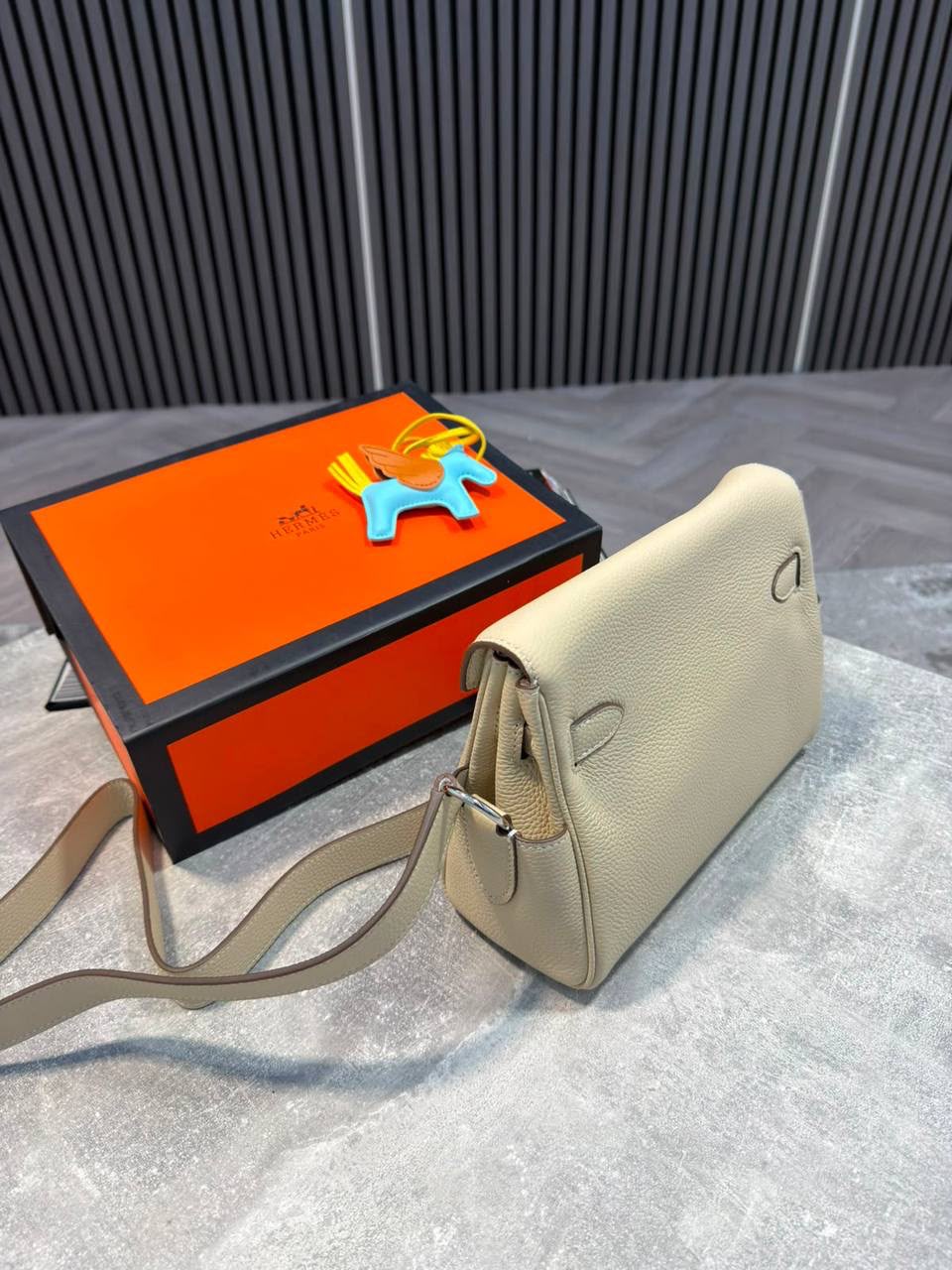 A light beige pebbled leather shoulder bag with a matching strap, displayed next to its signature orange and black Hermès box on a gray textured surface.
