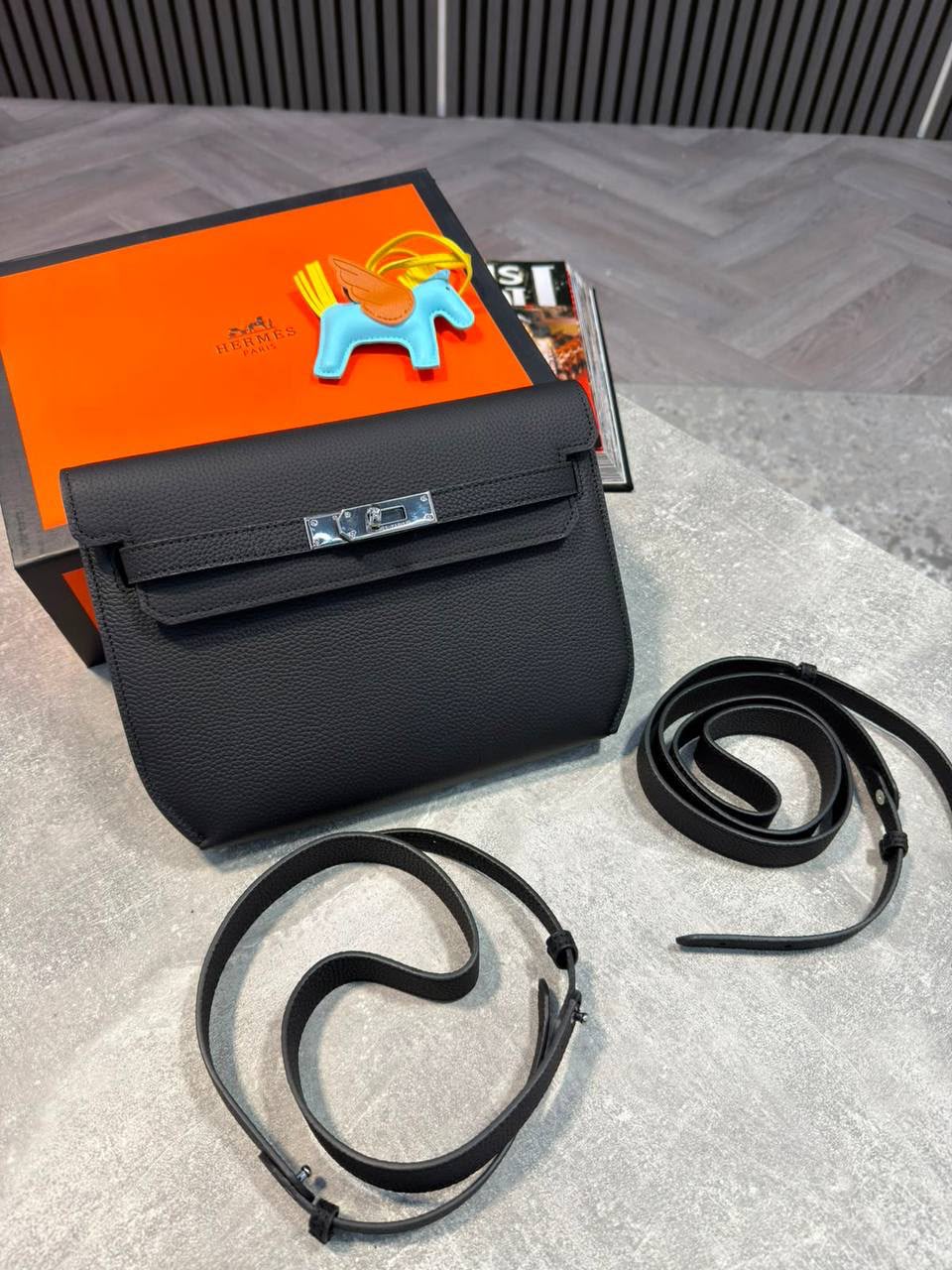 A black textured leather handbag with a silver clasp, shown with a detached long black strap on a light gray surface. An orange branded box and a small horse-shaped accessory are visible behind the bag.