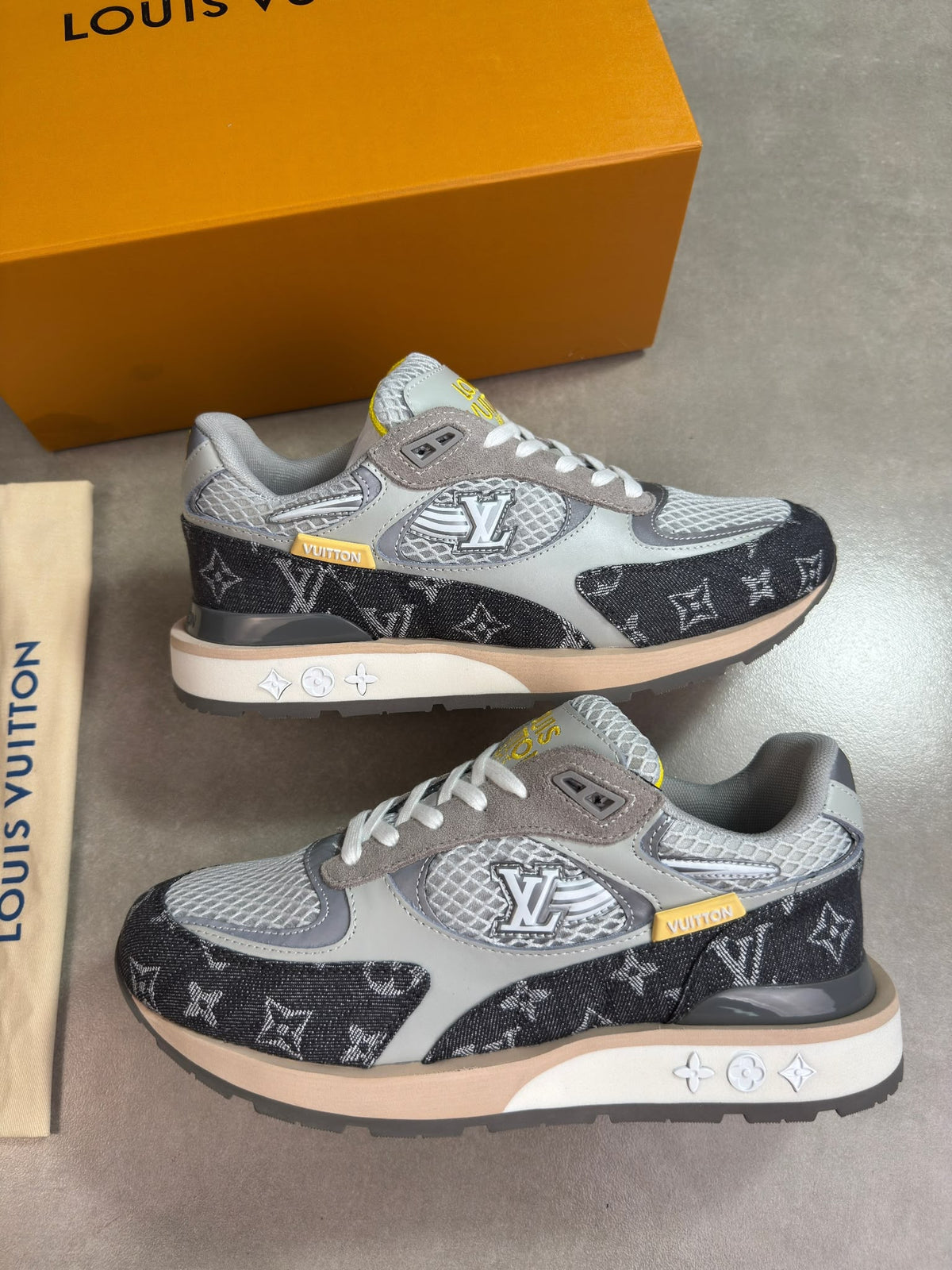 A pair of Louis Vuitton sneakers in gray, black, and beige with monogram patterns, displayed on a gray floor with a yellow Louis Vuitton box and a paper bag in the background.