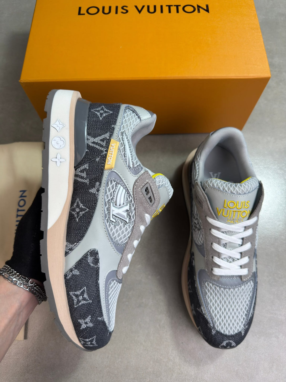 A pair of Louis Vuitton monogram sneakers shown with the orange Louis Vuitton box behind them. One shoe is held by a hand wearing a black glove, and the other sits on a gray floor.