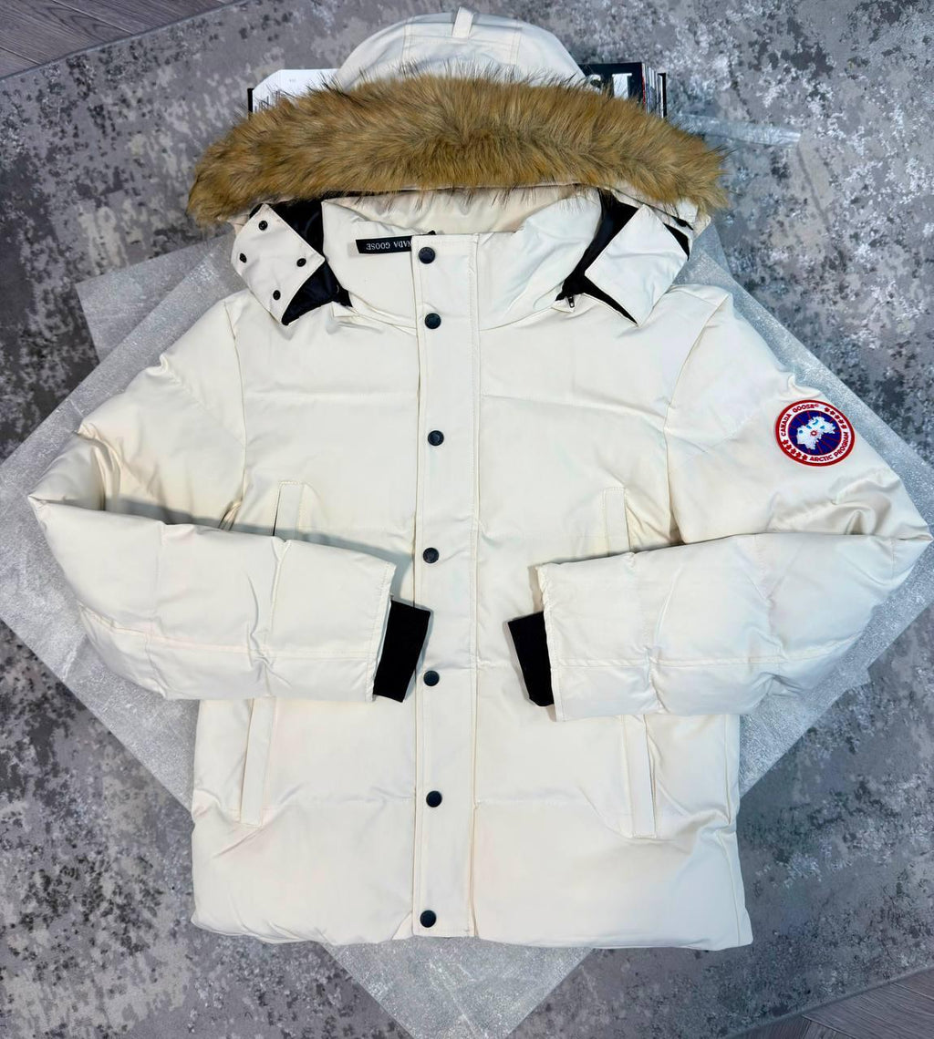 A cream-colored Canada Goose puffer parka jacket with a brown faux-fur trimmed hood, black snap buttons, and black cuff tabs, displayed flat on a gray textured surface.