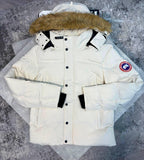A cream-colored Canada Goose puffer parka jacket with a brown faux-fur trimmed hood, black snap buttons, and black cuff tabs, displayed flat on a gray textured surface.