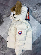A cream-colored Canada Goose puffer jacket with a brown faux-fur trimmed hood, laid flat on a gray textured surface with a magazine underneath.