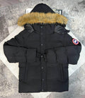 A black Canada Goose hooded puffer jacket with a tan faux-fur trimmed hood, laid flat on a light-colored geometric mat over a gray textured floor. The Canada Goose logo patch is visible on the left sleeve.