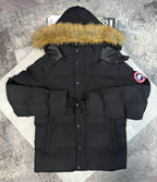 A black Canada Goose hooded puffer jacket with a tan faux-fur trimmed hood, laid flat on a light-colored geometric mat over a gray textured floor. The Canada Goose logo patch is visible on the left sleeve.