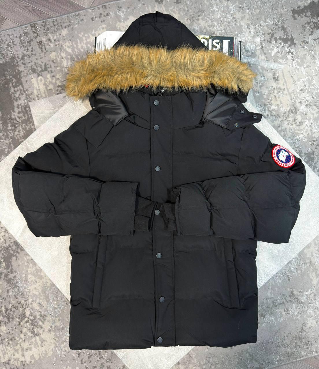 A black Canada Goose hooded puffer jacket with a tan faux-fur trimmed hood, laid flat on a light-colored geometric mat over a gray textured floor. The Canada Goose logo patch is visible on the left sleeve.