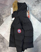 A black Canada Goose puffer coat with a tan faux-fur trim on the hood, laid flat on a light gray textured mat over a gray patterned floor. The Canada Goose logo patch is visible on the left front pocket.