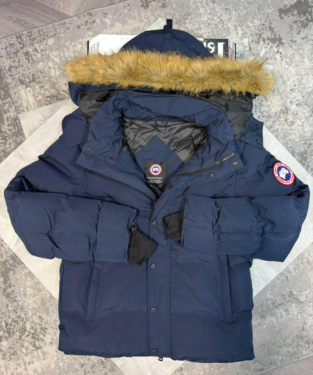 A navy blue Canada Goose puffer parka jacket with a tan faux-fur trimmed hood, laid flat on a light gray patterned surface. The jacket features the signature Canada Goose round patch on the left sleeve and a brand label on the inner neck.
