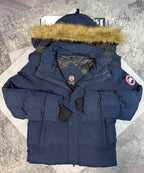A navy blue Canada Goose puffer parka jacket with a tan faux-fur trimmed hood, laid flat on a light gray patterned surface. The jacket features the signature Canada Goose round patch on the left sleeve and a brand label on the inner neck.
