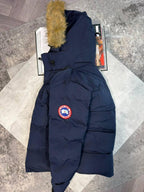 A navy blue Canada Goose puffer jacket with a tan faux-fur trimmed hood and the signature red, white, and blue logo patch on the chest, laid flat on a light gray stone surface.