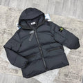A black Stone Island puffer jacket with a hood, laid flat on a gray textured rug over a wood-look floor. The Stone Island compass patch is visible on the left sleeve.