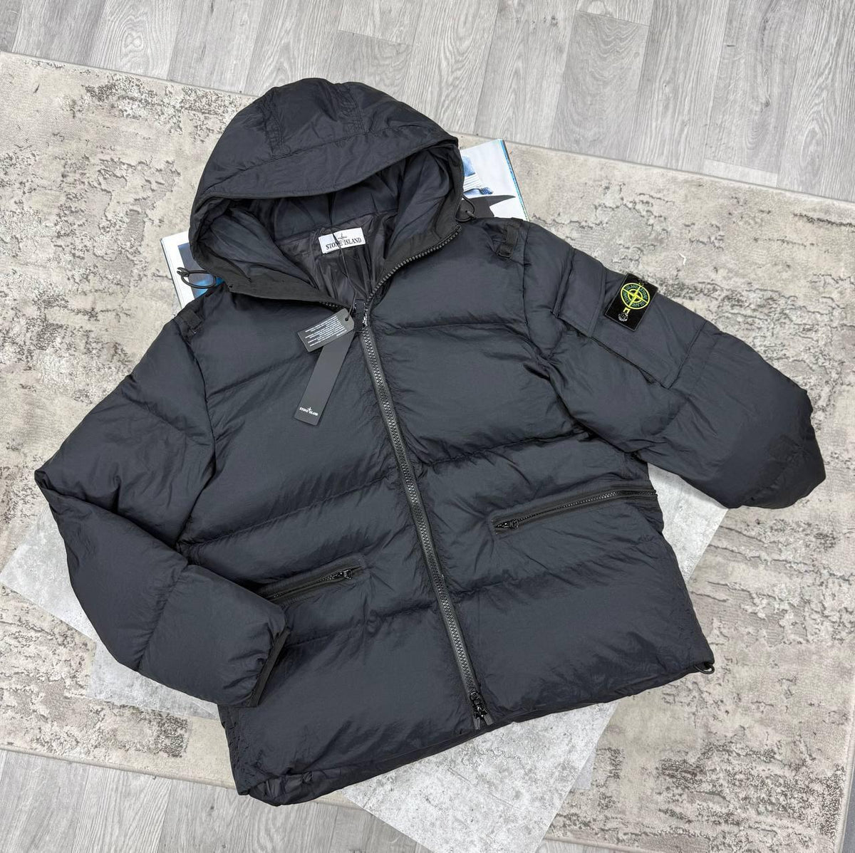 A black Stone Island puffer jacket with a hood, laid flat on a gray textured rug over a wood-look floor. The Stone Island compass patch is visible on the left sleeve.