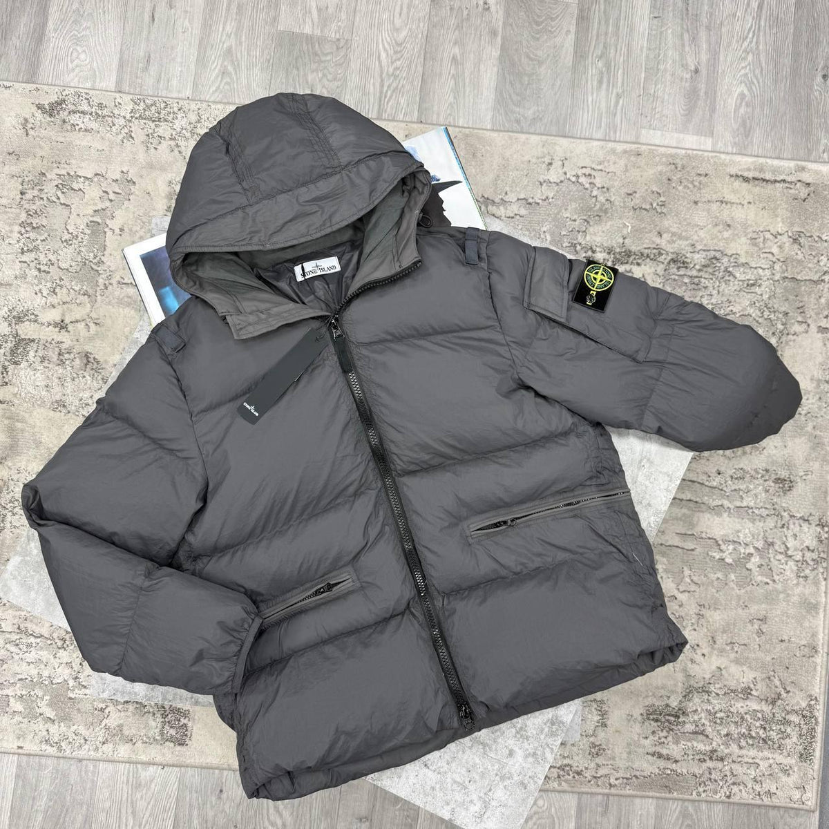 A dark gray Stone Island puffer jacket with the brand's compass logo patch on the sleeve, laid flat on a textured gray rug over a wood floor.