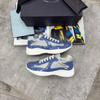 A pair of Prada low-top sneakers in blue patent leather with light gray mesh panels and white soles, displayed on a gray stone surface with a black Prada shoebox and magazine props.