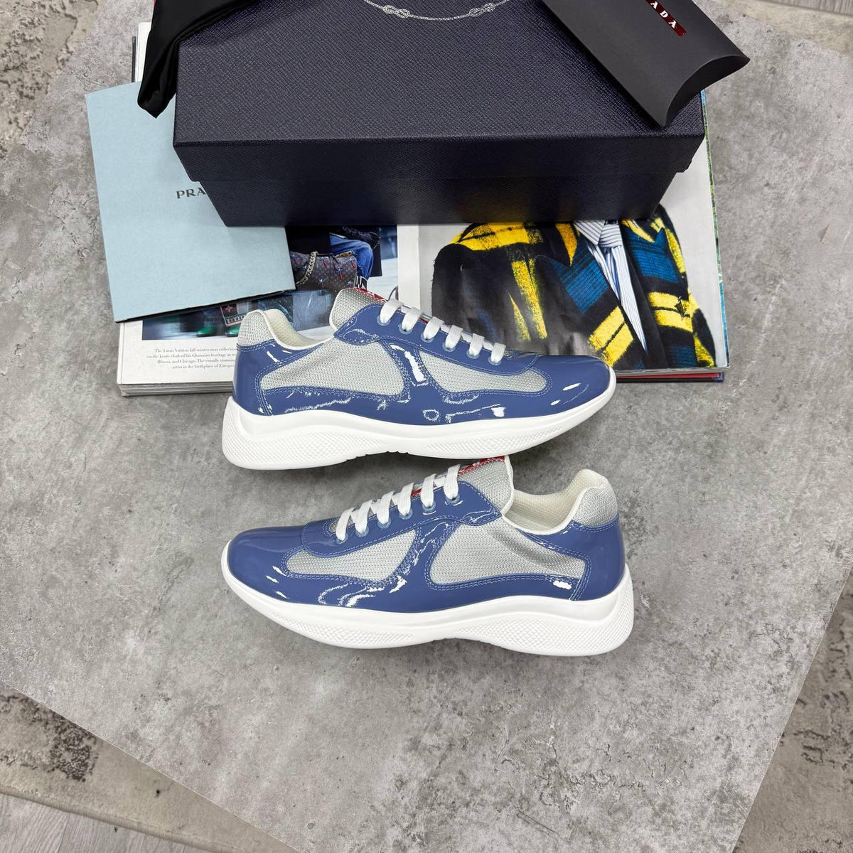 A pair of Prada low-top sneakers in blue patent leather with light gray mesh panels and white soles, displayed on a gray stone surface with a black Prada shoebox and magazine props.
