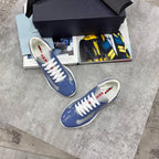 A pair of blue low-top sneakers with white laces and white soles, displayed on a gray stone surface next to an open magazine, a black shoebox, and a black accessory box.