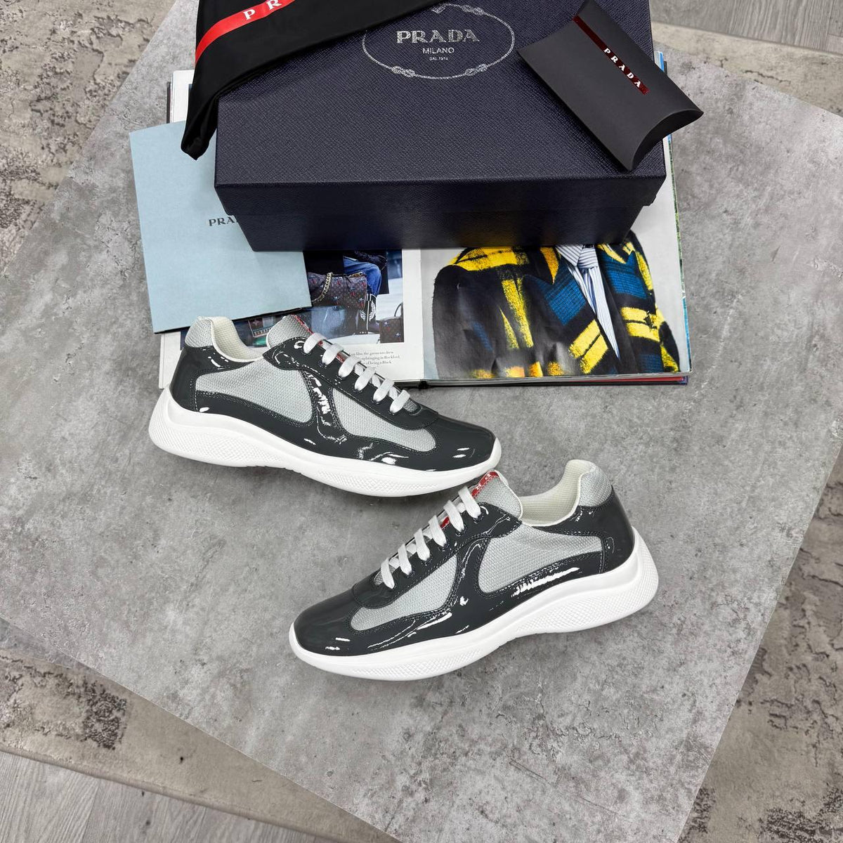 A pair of Prada low-top sneakers in glossy black and light gray with white soles, displayed next to their black Prada box, a branded belt, and a magazine on a gray stone surface.