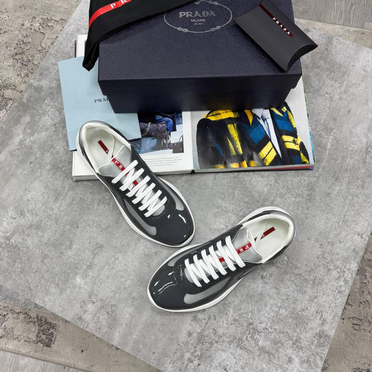 A pair of Prada low-top sneakers in glossy black and white with white laces and red accents, displayed next to their navy blue Prada box and a magazine on a gray stone surface.