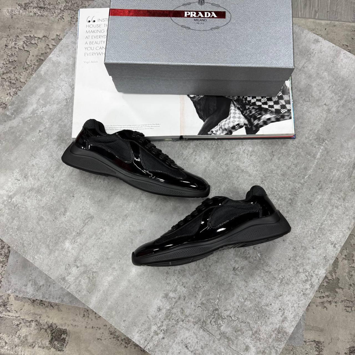 A pair of glossy black Prada sneakers displayed on a textured gray stone surface, with the gray Prada shoebox and a printed magazine or booklet placed behind them.