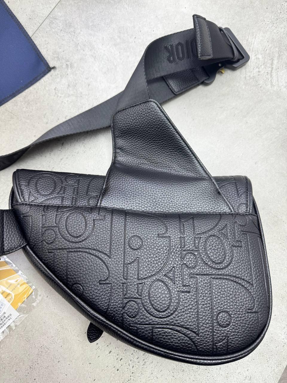 A black Dior saddle bag made of textured pebbled leather with an embossed monogram pattern, featuring a wide adjustable shoulder strap with a visible Dior logo.