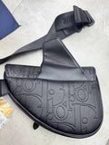A black Dior saddle bag made of textured pebbled leather with an embossed monogram pattern, featuring a wide adjustable shoulder strap with a visible Dior logo.
