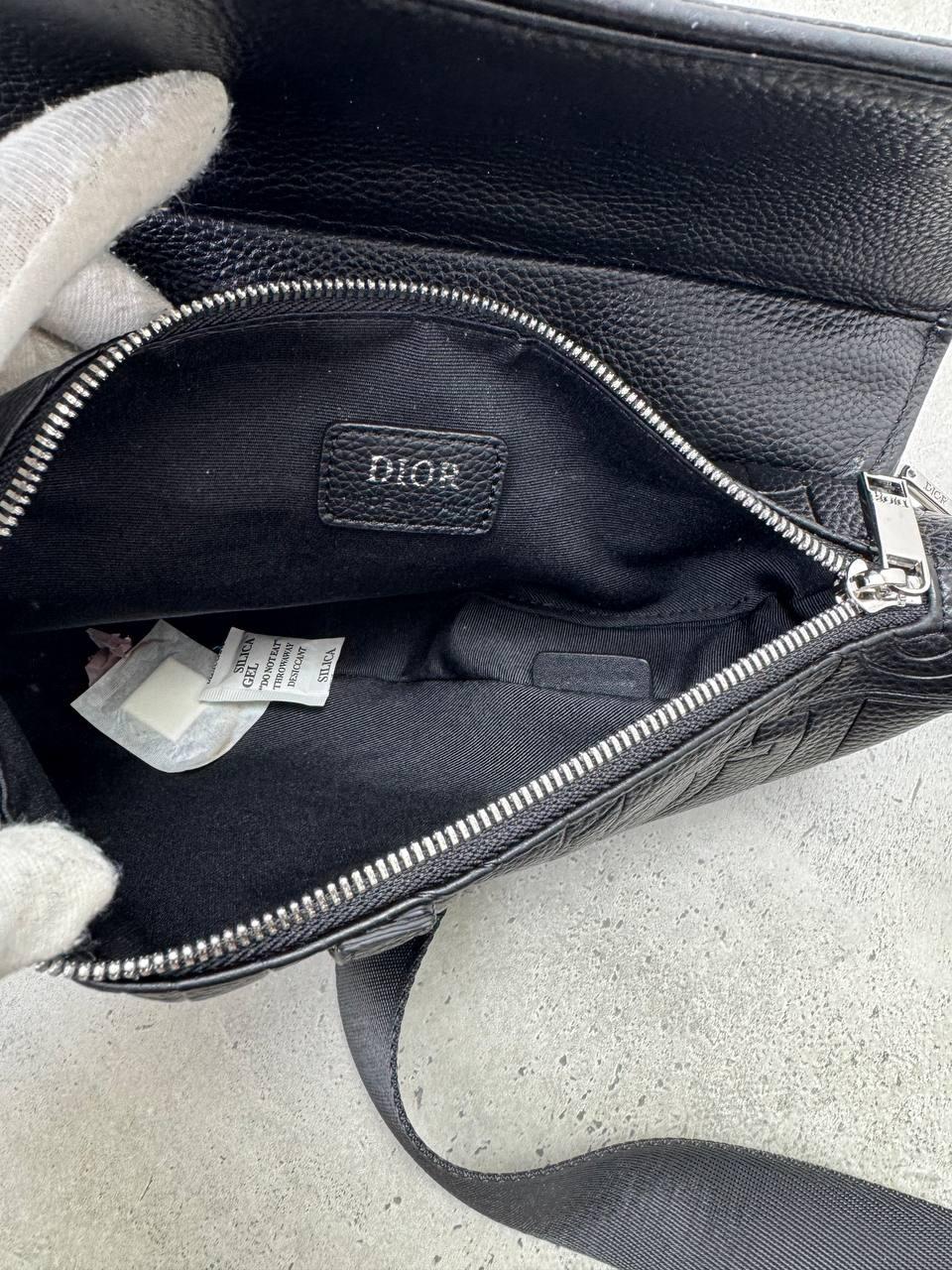 An open black leather Dior handbag shown from the inside, with a black fabric lining, silver zipper, and a Dior logo patch on the interior. A white-gloved hand is holding the bag open, and the product is photographed on a light gray concrete surface.