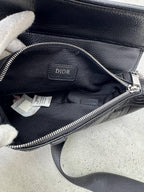 An open black leather Dior handbag shown from the inside, with a black fabric lining, silver zipper, and a Dior logo patch on the interior. A white-gloved hand is holding the bag open, and the product is photographed on a light gray concrete surface.