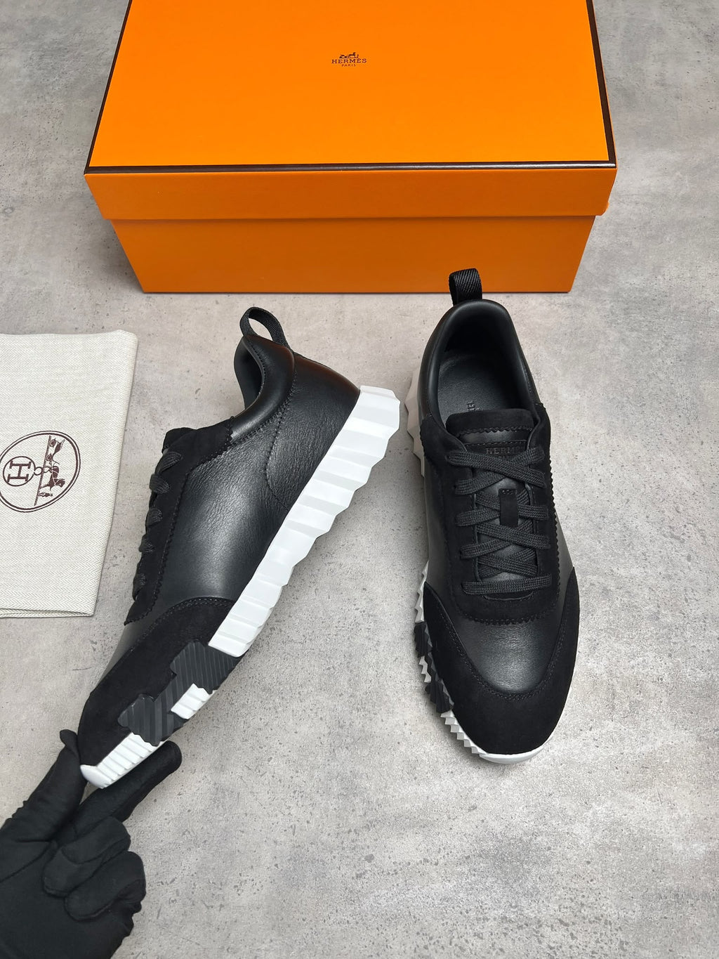 A pair of black leather low-top sneakers with chunky white sculptural soles, displayed next to their signature orange Hermès shoebox and a branded dust bag on a gray concrete surface.