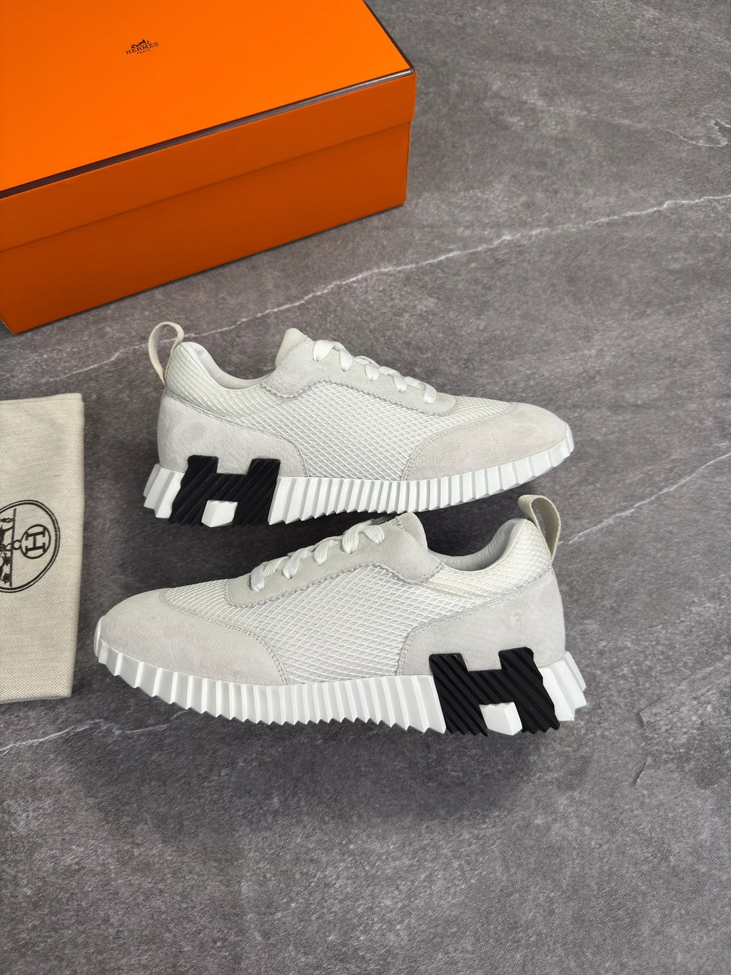 A pair of light gray and white knit sneakers with black woven 'H' side details, displayed next to an orange Hermès shoebox and a branded dust bag on a gray stone surface.