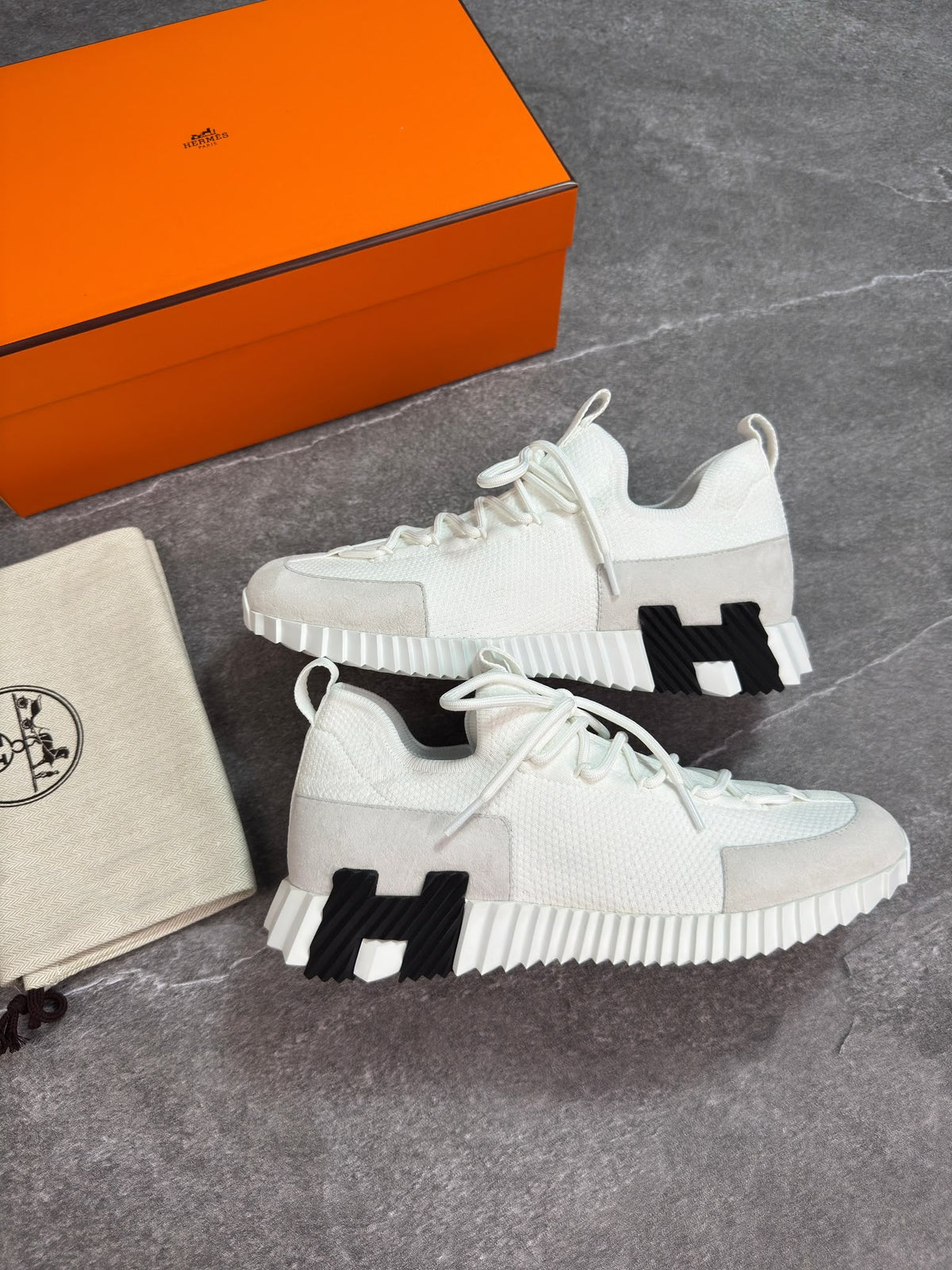 A pair of white and light gray knit sneakers with prominent black 'H' logos on the sides, displayed next to an orange Hermès box and a beige dust bag on a gray stone surface.