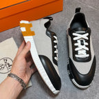 A pair of low-top sneakers in light gray and black with white laces and white soles, shown on a gray floor with an orange box and a branded dust bag. A hand is holding one shoe to display the side profile.