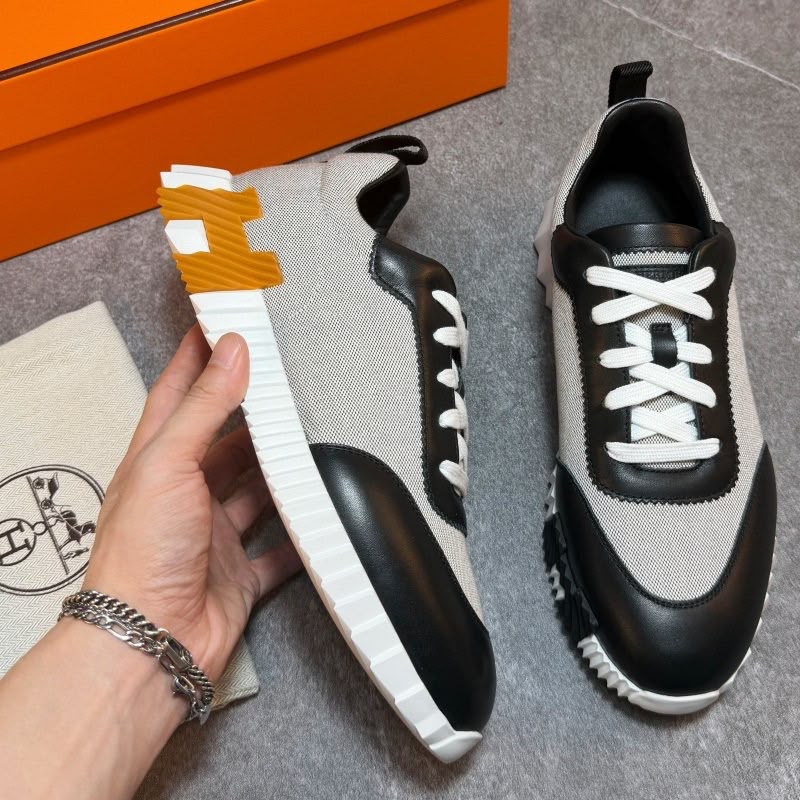 A pair of low-top sneakers in light gray and black with white laces and white soles, shown on a gray floor with an orange box and a branded dust bag. A hand is holding one shoe to display the side profile.