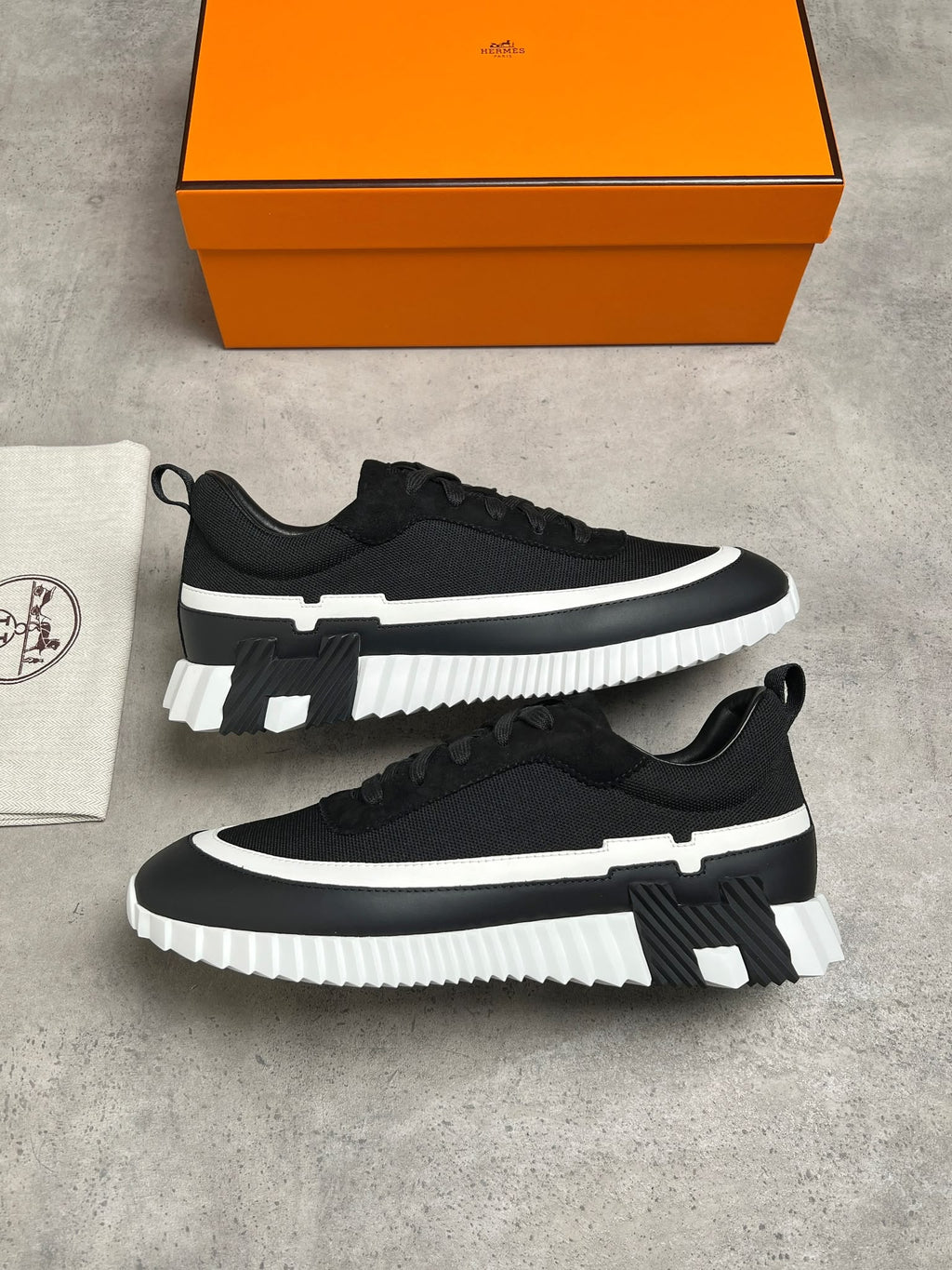 A pair of black low-top sneakers with chunky white platform soles, displayed on a gray concrete surface next to a white dust bag and an orange shoebox.