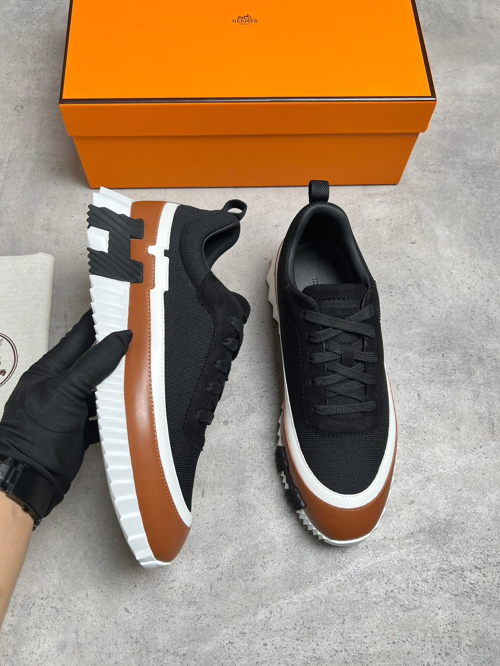 A pair of black low-top sneakers with brown and white chunky platform soles, shown on a gray concrete floor with an orange shoebox and a gloved hand holding one shoe.
