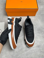 A pair of black low-top sneakers with brown and white chunky platform soles, shown on a gray concrete floor with an orange shoebox and a gloved hand holding one shoe.