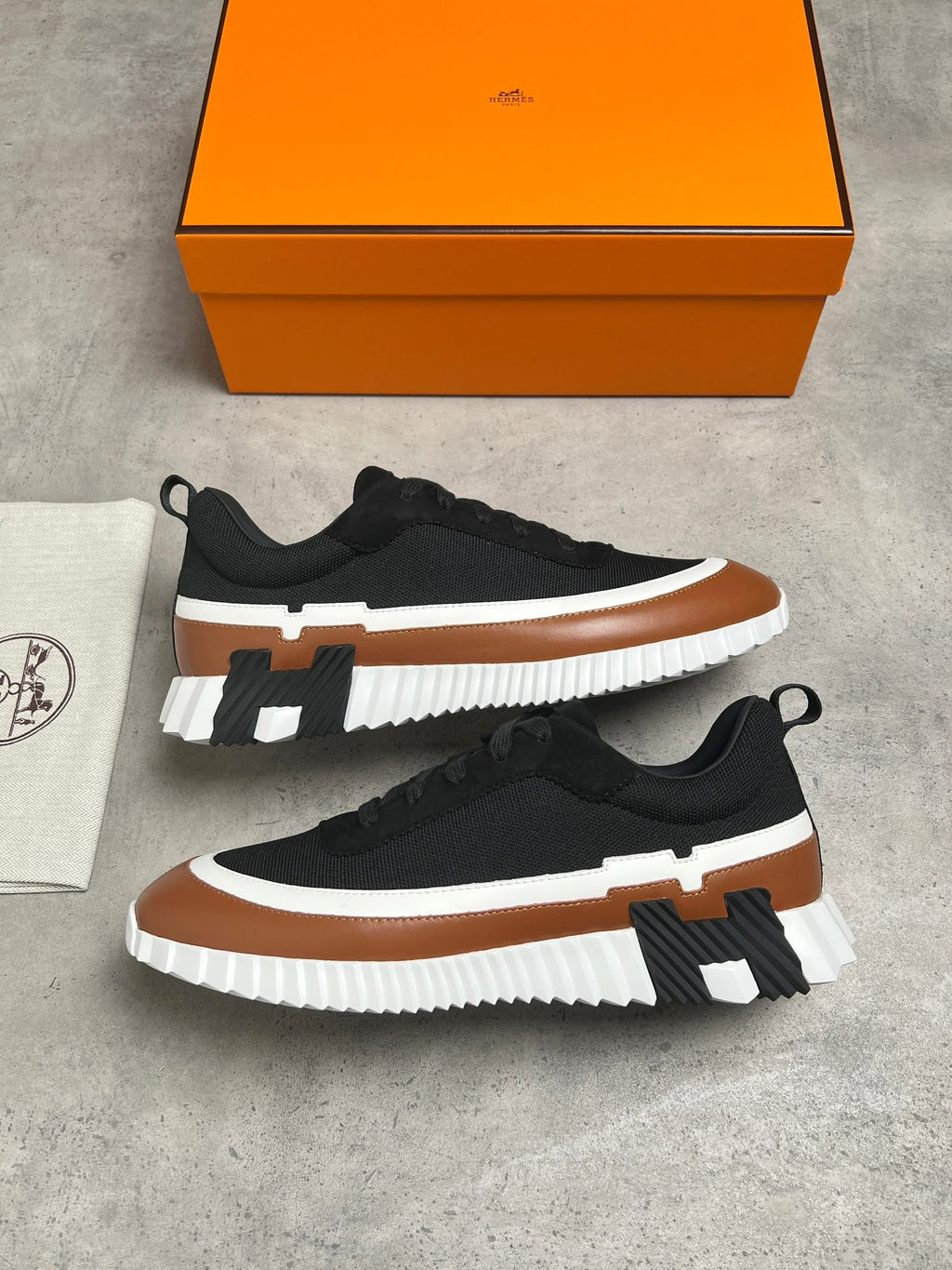A pair of black low-top sneakers with brown and white chunky soles and a black H-shaped accent, displayed on a gray concrete surface with an orange shoebox and a white dust bag.