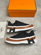 A pair of black low-top sneakers with brown and white chunky soles and a black H-shaped accent, displayed on a gray concrete surface with an orange shoebox and a white dust bag.