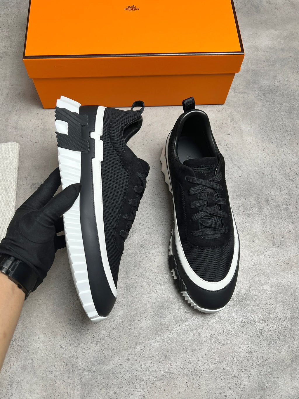 A pair of black low-top sneakers with chunky white and black soles, shown on a gray concrete floor with an orange shoebox in the background. One shoe is held by a gloved hand.